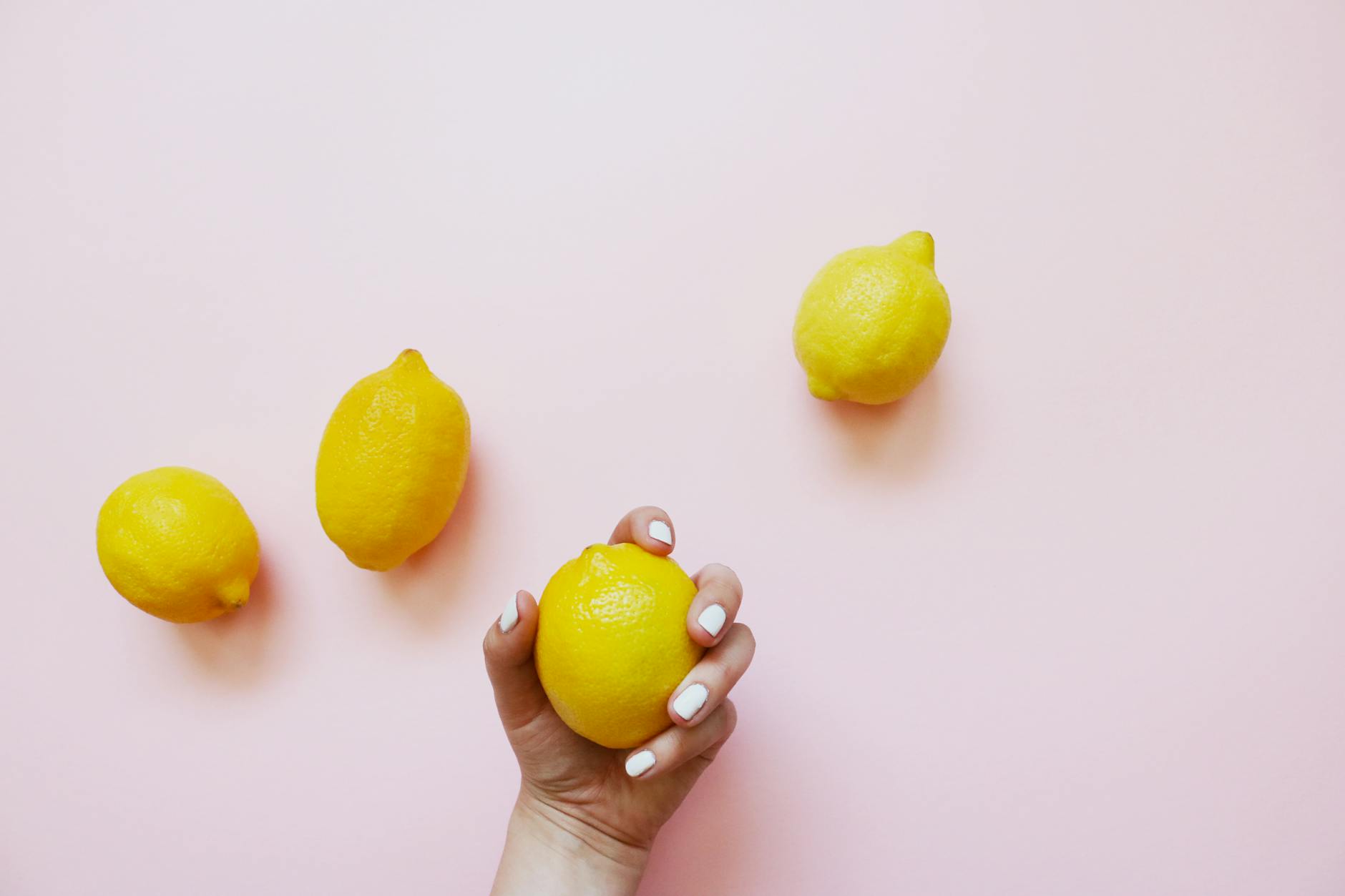 A hand holding a fresh lemon on a soft pink background with additional lemons nearby