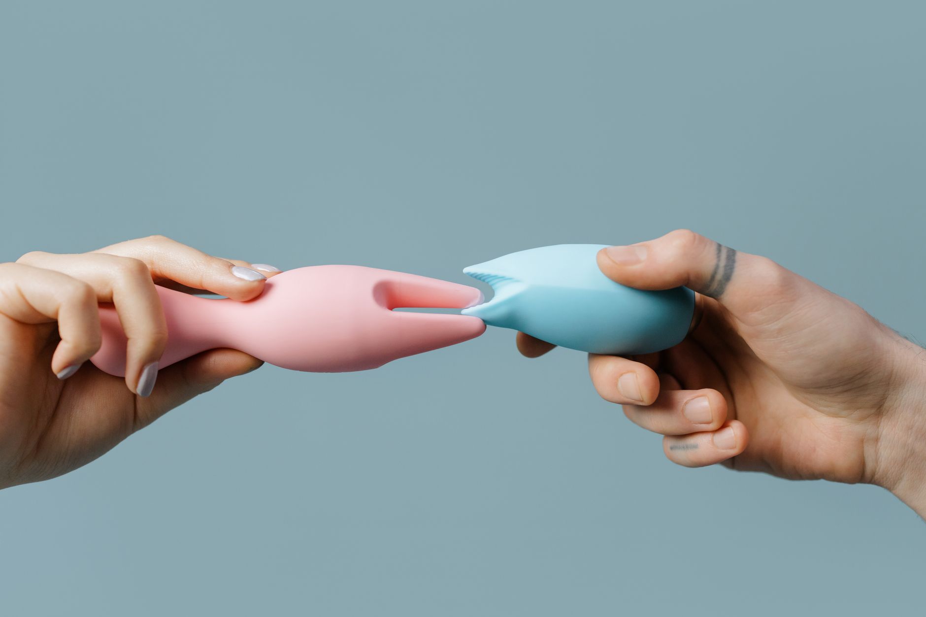 Hands holding pastel-colored silicone vibrators on a neutral background, representing shared pleasure and intimacy between partners