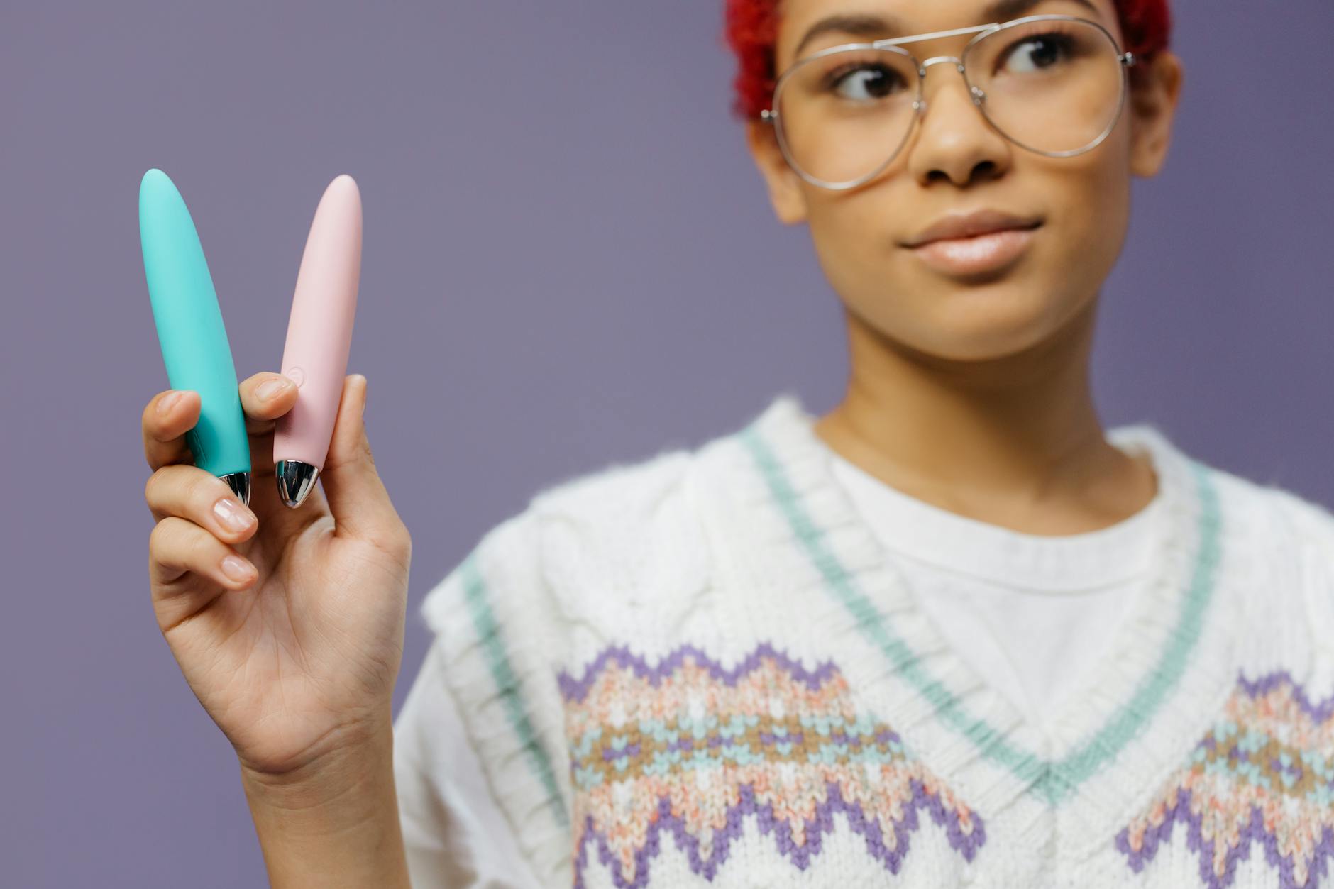 A person holding two colorful vibrators against a purple background, representing pleasure and sexual wellness.