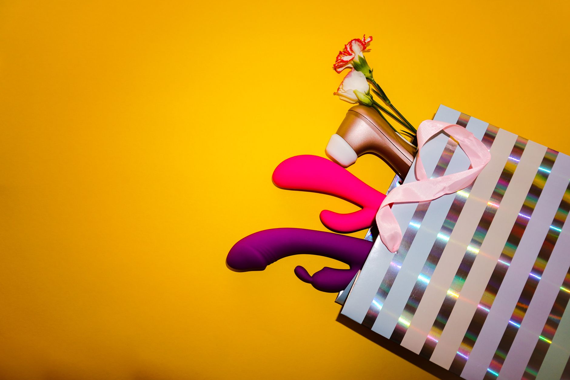 Colorful vibrators and wellness products arranged with fresh flowers in a gift bag against a bold yellow background.